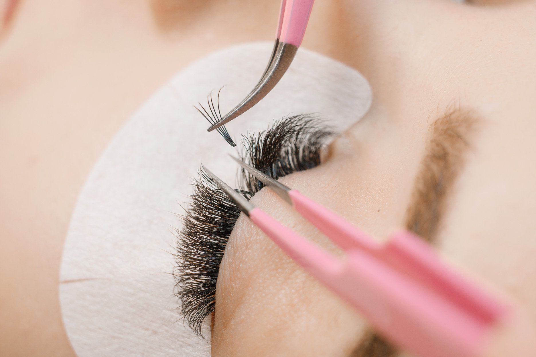 Eyelash extension procedure. Close up view of beautiful woman with long eyelashes. Stylist holding tweezers, tongs and making lengthening lashes for girl in a beauty salon. Beauty concept.